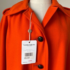 May include: Bright orange coat with a classic collar and large black buttons. The coat has a tag from Vestiaire Collective. The tag includes the text "Vestiaire Collective" and "CYNTHIA".