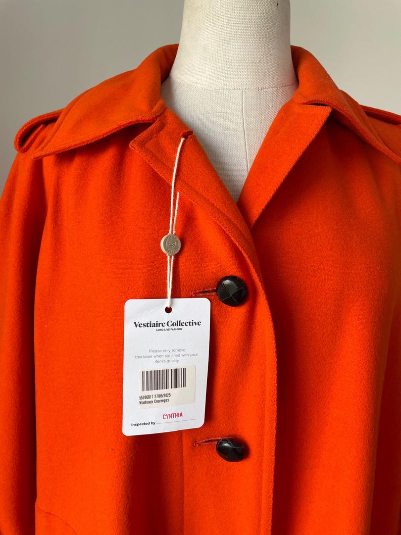 May include: Bright orange coat with a classic collar and large black buttons. The coat has a tag from Vestiaire Collective. The tag includes the text "Vestiaire Collective" and "CYNTHIA".