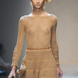 Könnte beinhalten: Beigefarbenes Häkelkleid mit langen Ärmeln und ausgestelltem Rock. Das Kleid hat einen braunen Gürtel und wird mit einem passenden Kopftuch und einer kleinen Clutch kombiniert. Das Kleid ist aus einem netzartigen Material gefertigt.