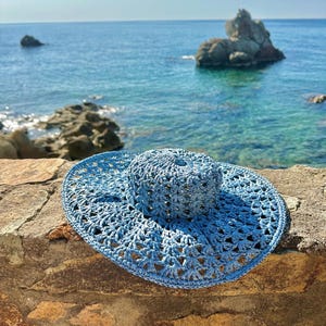 Puede incluir: Un sombrero de sol azul claro, de tejido abierto, con ala ancha. El sombrero está hecho de un material tejido y tiene una corona redondeada. El sombrero está sobre un muro de piedra con el mar de fondo.