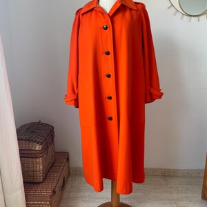 May include: A vibrant orange coat displayed on a wooden mannequin. The coat features a classic design with a collar, black buttons, and long sleeves. The coat is knee-length and appears to be made of a smooth fabric. The mannequin is on a wooden stand.