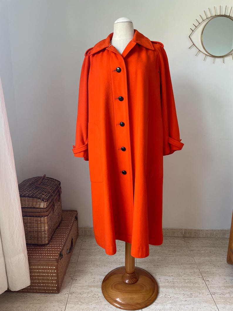 May include: A vibrant orange coat displayed on a wooden mannequin. The coat features a classic design with a collar, black buttons, and long sleeves. The coat is knee-length and appears to be made of a smooth fabric. The mannequin is on a wooden stand.