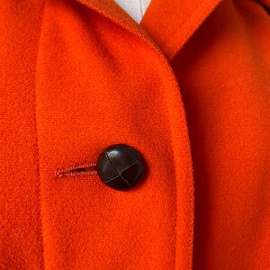 May include: Close-up of a vibrant orange coat with a dark brown button. The coat features a classic collar and a textured fabric. The button is round and appears to be leather or a similar material.