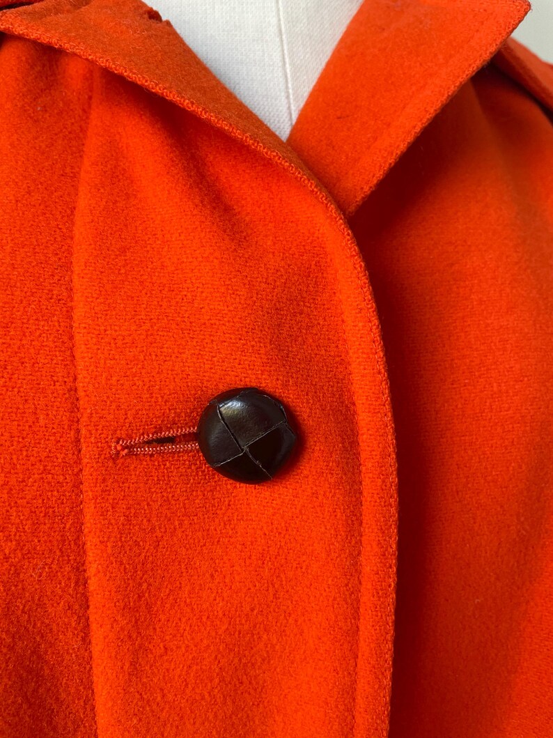 May include: Close-up of a vibrant orange coat with a dark brown button. The coat features a classic collar and a textured fabric. The button is round and appears to be leather or a similar material.