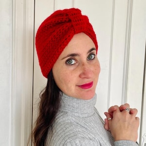 Puede incluir: Un turbante rojo tejido de punto. El gorro tiene una textura y un nudo en la parte superior. El gorro es de un solo color y parece cálido y cómodo. Un accesorio de moda para la cabeza.