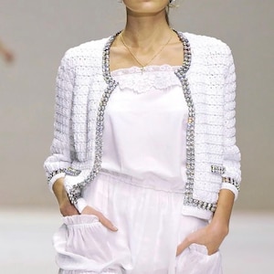 Puede incluir: Chaqueta blanca texturizada con ribete de joyas, combinada con un top y shorts blancos. Los shorts tienen un ribete de encaje. El conjunto forma parte de una colección de moda, mostrando una estética limpia y elegante.