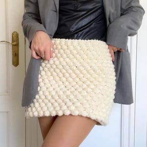 Puede incluir: Minifalda texturizada de color crema. La falda está hecha de un material grueso y tejido con un patrón de punto de burbuja. La falda se usa con una blazer gris y una parte superior negra.