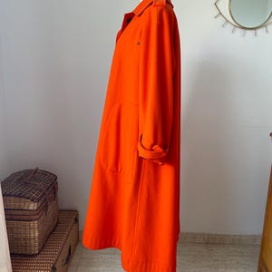 May include: A vibrant orange coat displayed on a wooden mannequin. The coat features a classic collar, large pockets, and rolled-up sleeves. The coat is knee-length and appears to be made of a soft, wool-like material. The coat is a bright, solid color.