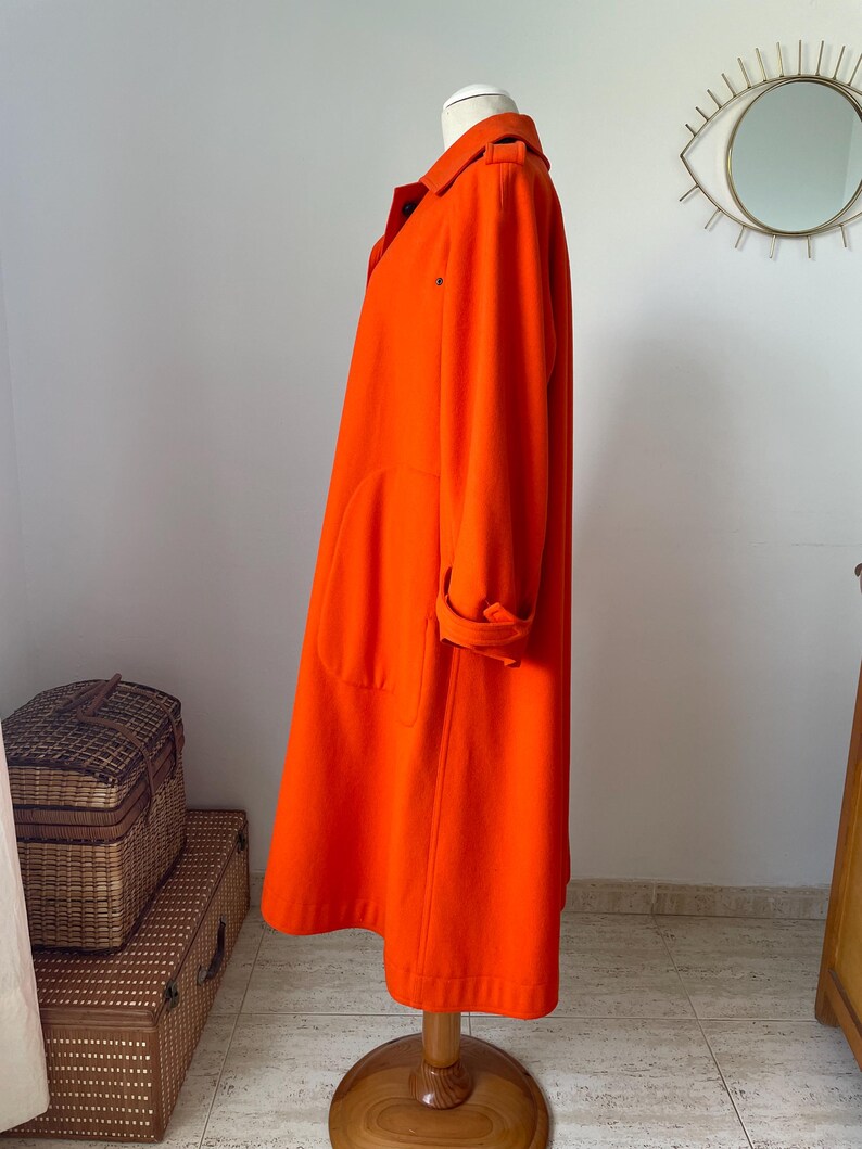 May include: A vibrant orange coat displayed on a wooden mannequin. The coat features a classic collar, large pockets, and rolled-up sleeves. The coat is knee-length and appears to be made of a soft, wool-like material. The coat is a bright, solid color.