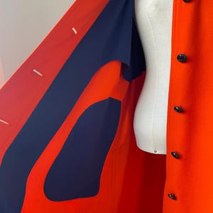 May include: A vibrant orange coat with a navy blue lining and black buttons. The coat is displayed on a mannequin, showcasing its tailored design and contrasting color scheme. The coat's interior is visible, highlighting the navy blue fabric.