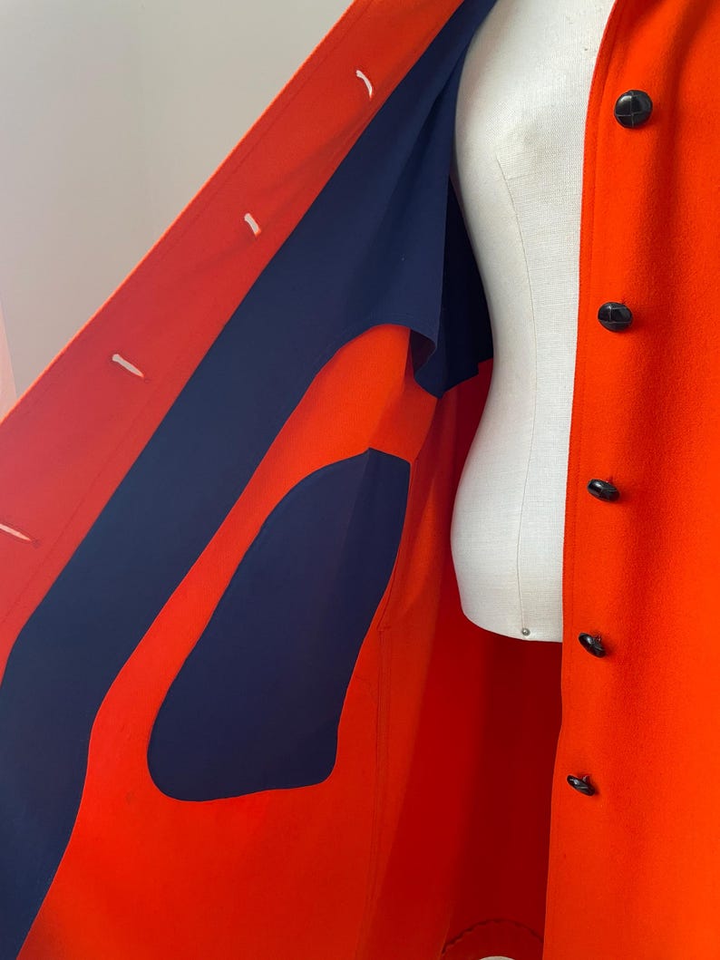 May include: A vibrant orange coat with a navy blue lining and black buttons. The coat is displayed on a mannequin, showcasing its tailored design and contrasting color scheme. The coat's interior is visible, highlighting the navy blue fabric.