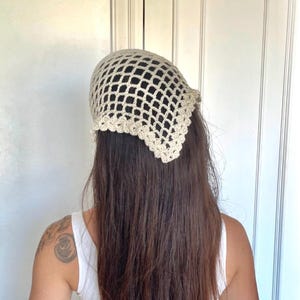 May include: A beige crochet headscarf with a net-like design. The head covering is worn on the head and has a scalloped edge. The headscarf is made of a light-colored yarn and is a handmade item.