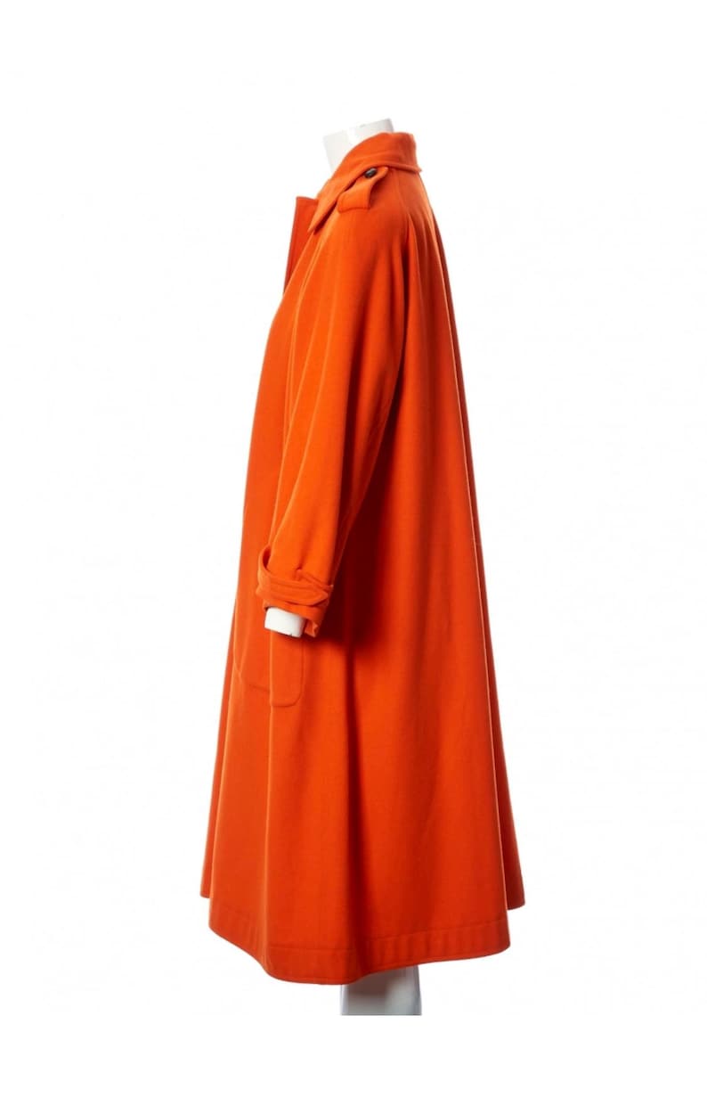 May include: A vibrant orange coat with a classic design. The coat features a collared neckline, long sleeves, and a knee-length hem. The coat is displayed on a mannequin, showcasing its tailored fit and overall style. The coat has a button closure and pockets.