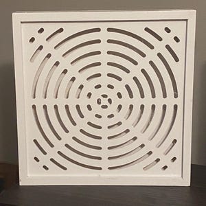 Può includere: Un coperchio di ventilazione quadrato in legno bianco con un design circolare concentrico. Il design presenta una serie di fessure curve che si irradiano dal centro, creando un motivo. Il telaio ha un design semplice e pulito.