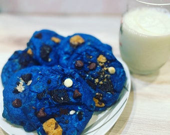 May include: A plate of blue cookies with chocolate chips, white chocolate chips, and cookie dough pieces. The cookies are on a white plate with a glass of milk in the background.
