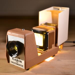 May include: A retro brown and beige slide projector, labelled "CABIN for INSTAMATIC". The projector is illuminated internally, with a black power cable extending from the rear. The lens is black, and the design is vintage.