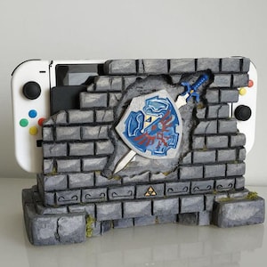 Puede incluir: Una pared de piedra gris con una sección rota que revela un escudo azul y rojo con una espada que sobresale. La pared es un soporte para una consola Nintendo Switch blanca.