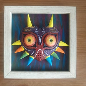 Puede incluir: Una escultura de madera enmarcada de la Máscara de Majora de la serie de videojuegos The Legend of Zelda. La máscara está pintada en tonos de rojo, naranja, amarillo, verde y azul, con detalles intrincados y puntas afiladas.