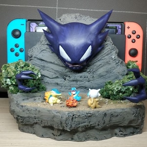Könnte beinhalten: Ein 3D-gedrucktes Diorama mit einer lila und schwarzen Gengar-Pokémon-Figur, einer Nintendo Switch-Konsole und mehreren kleineren Pokémon-Figuren. Das Diorama ist vor einem grauen Fels Hintergrund mit grünem Laub platziert.