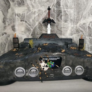 Puede incluir: Una consola Nintendo 64 negra con una pintura personalizada que se asemeja a una mazmorra oscura y cubierta de vegetación. La consola tiene una Espada Maestra de The Legend of Zelda: Ocarina of Time en la parte superior y una figura de araña en la parte delantera.