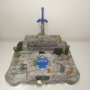 Könnte beinhalten: Eine graue Nintendo 64 Konsole mit einer steinartigen Textur. Die Konsole ist mit einem blauen und silbernen Master Sword, einem blauen und silbernen Hylian Shield und einer kleinen, grauen Spinnenfigur verziert. Die Konsole ist von kleinen, braunen Blättern und grünem Moos umgeben.