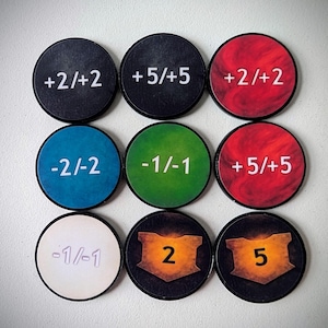 May include: Nine circular game tokens with various colors and markings. The tokens display values such as +2/+2, +5/+5, -2/-2, and -1/-1. Two tokens feature the numbers 2 and 5 within a shield design. The tokens are likely used in a board game.