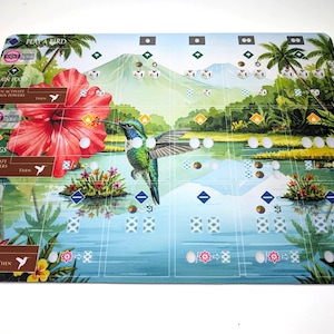 Op de afbeelding: Een kleurrijke spelmat met een kolibrie, een grote rode hibiscusbloem en een tropisch landschap met bergen en water. De mat bevat spelinstructies en ruimtes voor spelstukken.