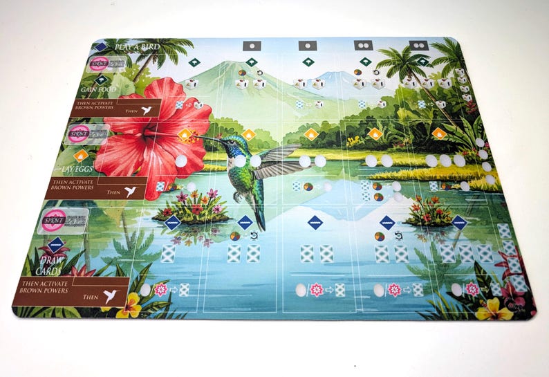 Op de afbeelding: Een kleurrijke spelmat met een kolibrie, een grote rode hibiscusbloem en een tropisch landschap met bergen en water. De mat bevat spelinstructies en ruimtes voor spelstukken.