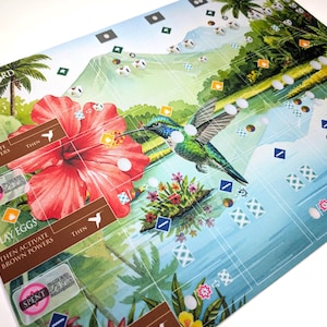 Op de afbeelding: Een kleurrijk bordspelcomponent met een kolibrie, een grote rode hibiscusbloem en tropische landschappen. Het spelbord bevat tekst zoals "PLAY A BIRD", "GAIN FOOD" en "LAY EGGS". Het spelbord ligt op een wit oppervlak.