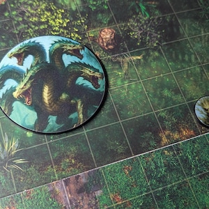 K&ouml;nnte beinhalten: Zwei runde Spielmarken mit Fantasy-Kreaturen. Eine Marke zeigt eine mehrk&ouml;pfige Hydra mit offenem Maul, die andere einen Waldgeist. Die Marken liegen auf einem gr&uuml;nen Spielbrett mit Rastermuster.