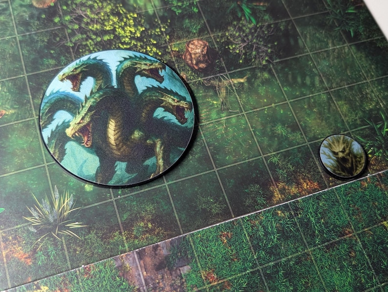 K&ouml;nnte beinhalten: Zwei runde Spielmarken mit Fantasy-Kreaturen. Eine Marke zeigt eine mehrk&ouml;pfige Hydra mit offenem Maul, die andere einen Waldgeist. Die Marken liegen auf einem gr&uuml;nen Spielbrett mit Rastermuster.