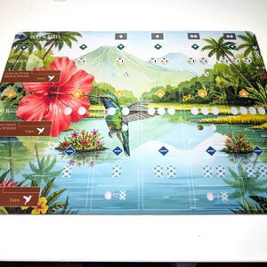 Op de afbeelding: Een rechthoekig spelbord met een levendige tropische sc&egrave;ne, met een kolibrie, een grote rode hibiscusbloem en een berg op de achtergrond. Het bord bevat tekst zoals "Play a Bird" en "Draw Cards".