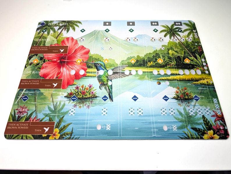 Op de afbeelding: Een rechthoekig spelbord met een levendige tropische sc&egrave;ne, met een kolibrie, een grote rode hibiscusbloem en een berg op de achtergrond. Het bord bevat tekst zoals "Play a Bird" en "Draw Cards".