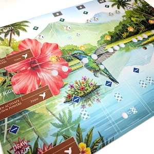 Op de afbeelding: Een kleurrijke spelmat met een kolibrie in de buurt van een grote rode hibiscusbloem. De mat heeft een tropisch landschapsontwerp met palmbomen, water en bergen. Tekst op de mat omvat "PLAY A BIRD", "GAIN FOOD" en "LAY EGGS".