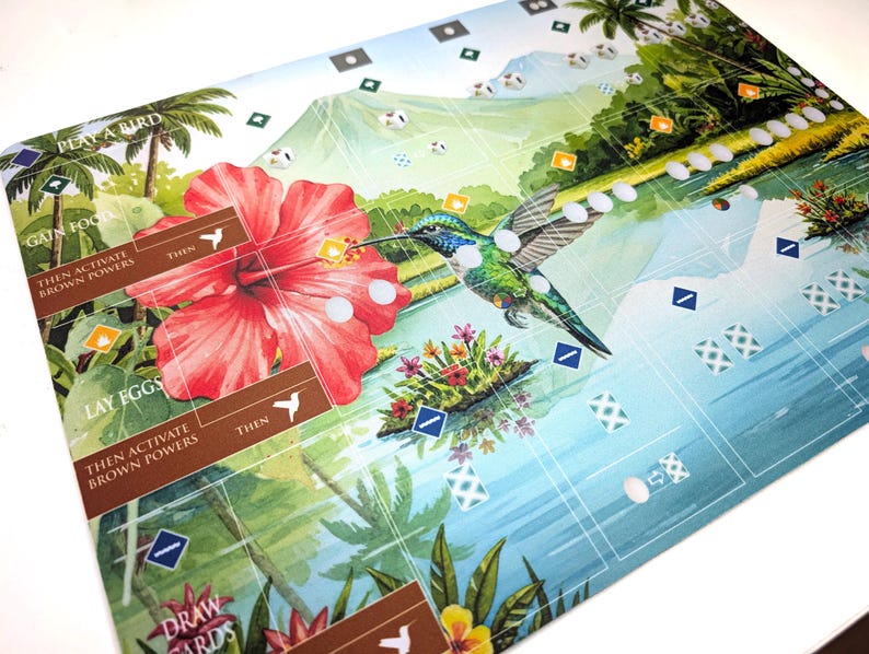 Op de afbeelding: Een kleurrijke spelmat met een kolibrie in de buurt van een grote rode hibiscusbloem. De mat heeft een tropisch landschapsontwerp met palmbomen, water en bergen. Tekst op de mat omvat "PLAY A BIRD", "GAIN FOOD" en "LAY EGGS".