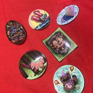 Op de afbeelding: Zes ronde stickers met verschillende cartoon-dierontwerpen. De stickers tonen een blauw en groen wezen met een boog en pijl, een bruin en wit wezen met een rode stok, een bruin en wit wezen met een paarse hoed, een blauw en paars wezen met vleugels, twee bruine en witte wezens die knuffelen, en een groen wezen met een rood en wit bootje.