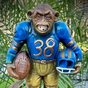 May include: A quirky statue of a monkey dressed in a football uniform. The monkey is wearing a blue jersey with gold detailing and the number 38. It holds a football and a helmet, both blue. The statue is brightly coloured and stands on a green base.