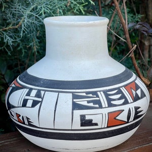 Puede incluir: Un jarrón de cerámica blanco con cuerpo redondeado y cuello acampanado. El jarrón presenta un diseño geométrico en blanco y negro con detalles en rojo. El jarrón es probablemente hecho a mano y tiene una estética tradicional del suroeste.