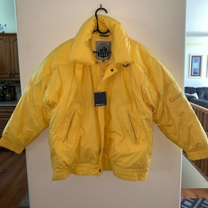 Op de afbeelding: Felge gele winterjas met een hoge kraag en meerdere zakken met ritssluiting. De jas heeft een label met de tekst "Down Is..." en "DASH ADVENTURE". De jas hangt aan een zwarte hanger.