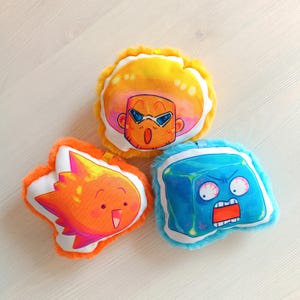 Puede incluir: Tres peluches con caras de personajes de dibujos animados. Uno es amarillo con un personaje con gafas de sol, otro es naranja y con forma de llama, y el tercero es azul y con forma de cubo. Cada juguete tiene un borde esponjoso.