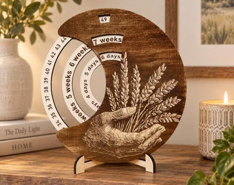 May include: A circular wooden pregnancy countdown calendar with a dark brown finish. The front features an engraved hand holding wheat stalks. It has rotating inner discs displaying weeks and days, with text including 7 weeks, 6 days, and numbers 34 through 49.