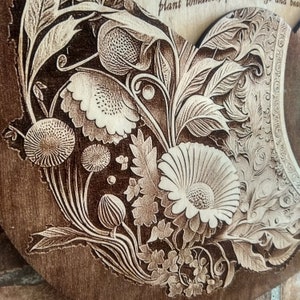 Peut inclure: Un gros plan d'une plaque en bois avec un motif floral d&eacute;taill&eacute; grav&eacute; &agrave; la surface. Le motif pr&eacute;sente une vari&eacute;t&eacute; de fleurs, de feuilles et de tiges, toutes finement grav&eacute;es dans une couleur brun clair sur un fond brun fonc&eacute;.