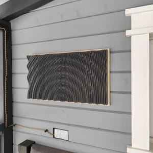 May include: A dark brown, textured wall art piece with a wave-like pattern. The artwork is rectangular and framed with a light-colored wood. The piece is mounted on a gray, horizontal-planked wall.