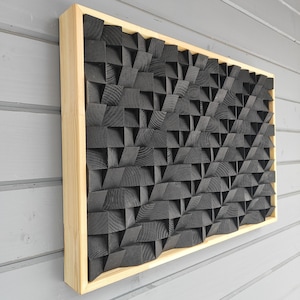 以下が含まれることがあります： 天然木のフレームが付いた長方形の木製ウォールアート。内部は、黒い幾何学的な木製ブロックのテクスチャパターンで、3D効果を生み出しています。アートワークは灰色の壁に取り付けられています。