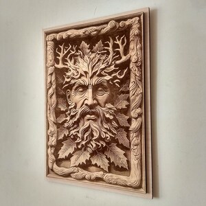 Puede incluir: Una talla de madera para la pared que representa al Hombre Verde, una figura m&iacute;tica con un rostro rodeado de hojas y ramas. La talla est&aacute; enmarcada con un borde decorativo.