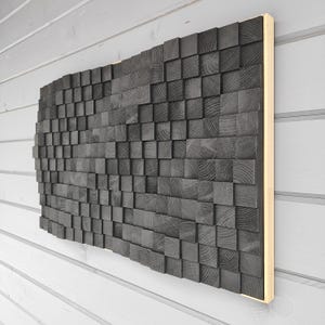 Könnte beinhalten: Ein großes, rechteckiges Wandkunstwerk, das aus zahlreichen dunkelgrauen Holzblöcken besteht. Die Blöcke sind in einem Mosaikmuster angeordnet und erzeugen eine strukturierte Oberfläche. Das Kunstwerk ist auf einem hellfarbigen Holzrahmen montiert.