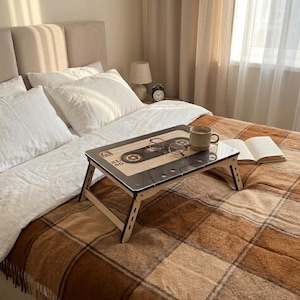 May include: A rectangular wooden bed tray featuring a graphic print of a vintage cassette tape with the text XL 90. The tray has foldable legs and holds a ceramic mug and a pair of wire-rimmed glasses, positioned on a brown plaid blanket.