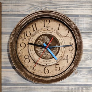 Op de afbeelding: Een houten klok met een verweerde bruine afwerking. De wijzerplaat van de klok heeft een golfontwerp en de woorden "Hoogwater", "Laagwater", "Halve vloed" en "Voor hoogwater" en "Na hoogwater". De wijzers van de klok zijn zwart, rood en blauw.
