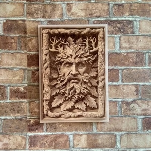 Puede incluir: Un adorno de pared de madera con un relieve tallado del Hombre Verde, una figura m&iacute;tica con rostro humano rodeado de hojas y ramas. La talla es detallada e intrincada, con un acabado marr&oacute;n r&uacute;stico.