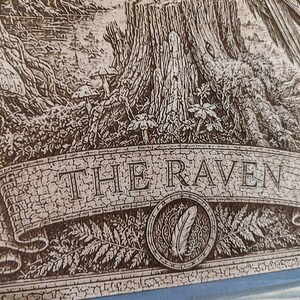 May include: A detailed wood-engraved illustration featuring a forest scene with a tree stump, mushrooms, and ferns. A prominent banner across the bottom displays the text THE RAVEN in capital letters, with a circular emblem containing a feather.